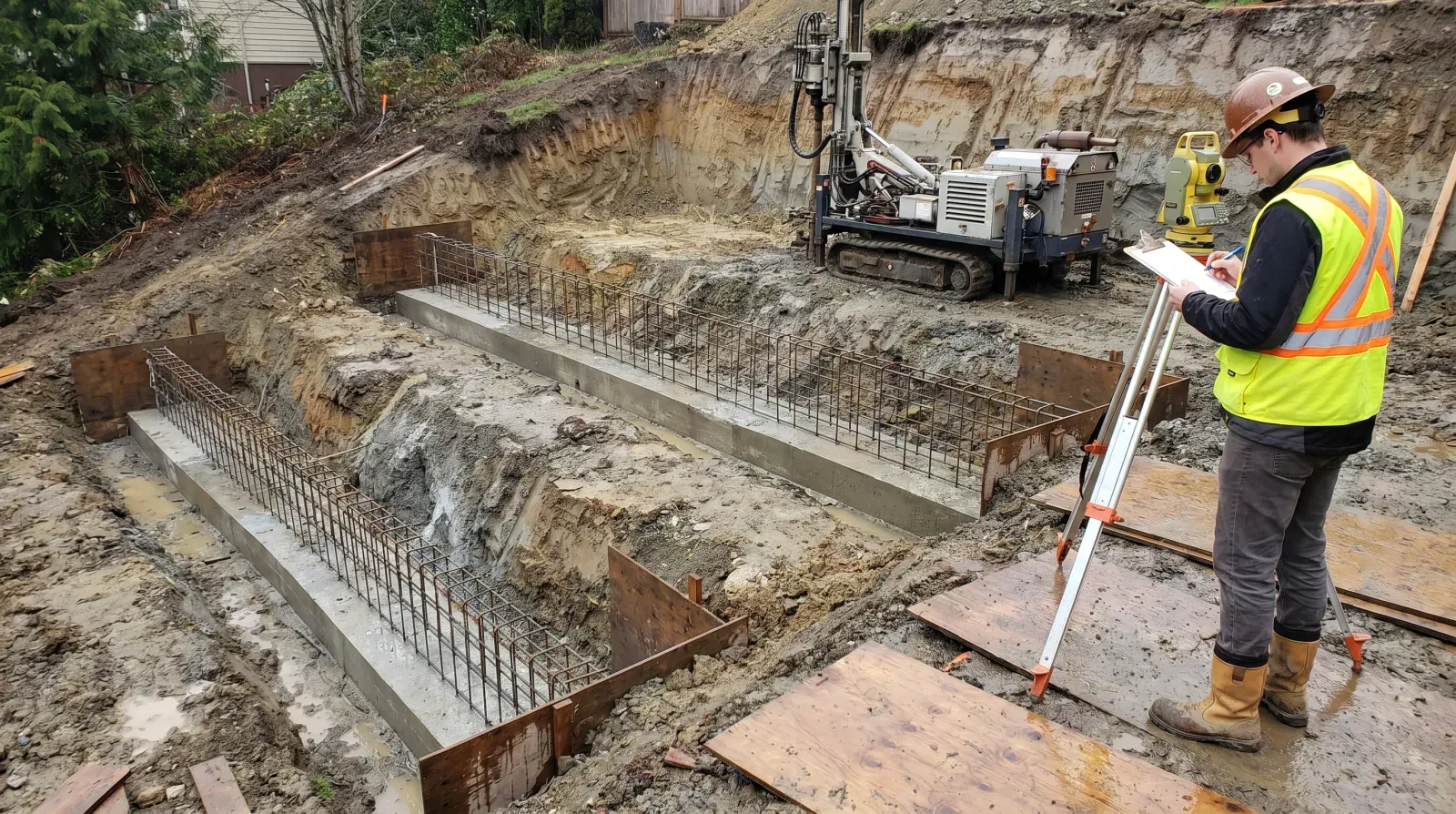 A stepped concrete strip footing with tied rebar cages descending a Burnaby hillside excavation, with a geotechnical engineer recording observations and a drilled-pile rig in the background.
